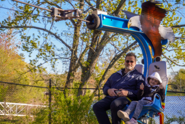 An adult and child sitting on the Soaring Eagle Zip Ride. Both are smiling and child is holding two thumbs up.